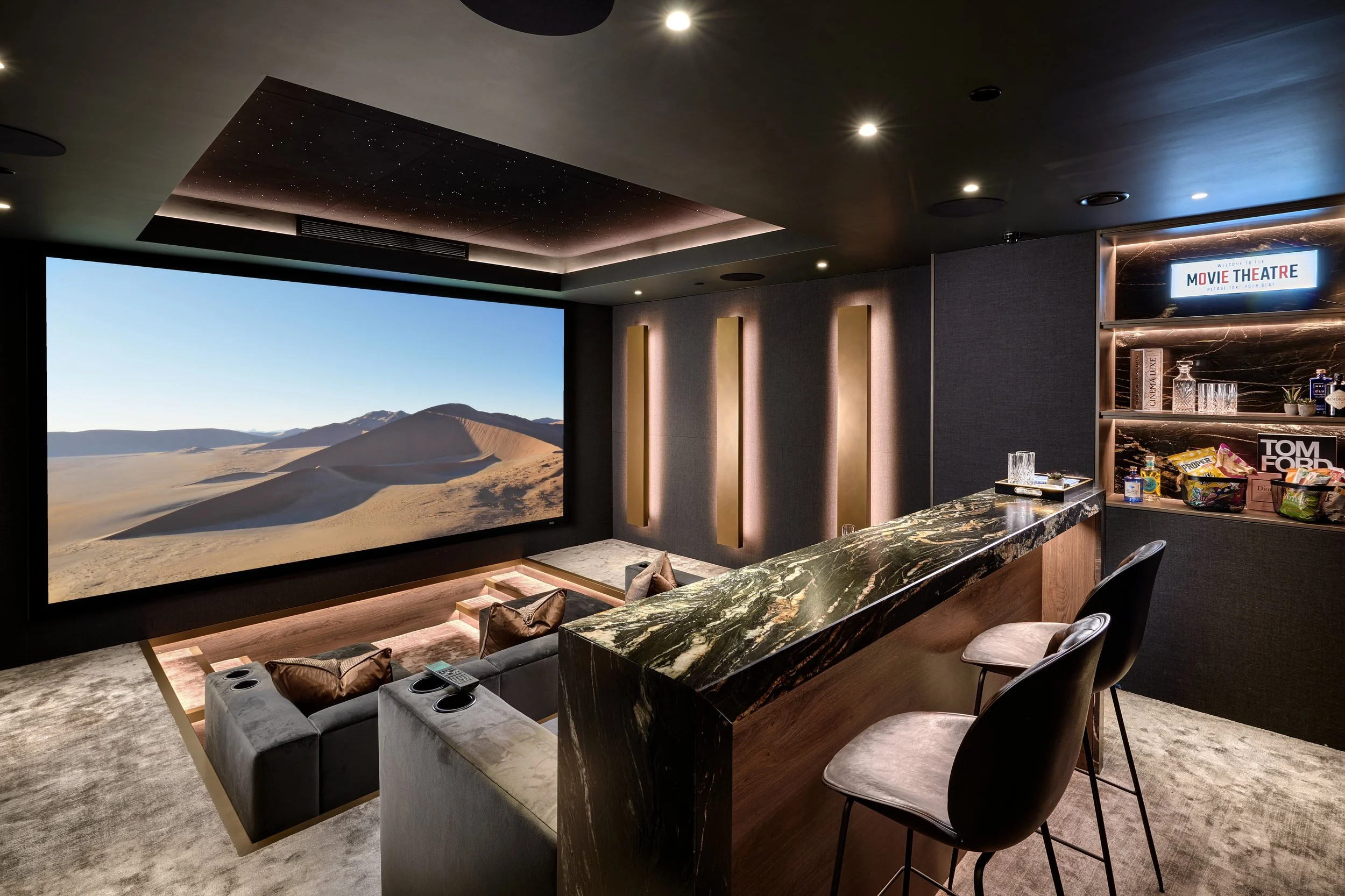 Luxury home theater room with a large screen displaying desert landscape, plush seating, a bar area with snacks and drinks, and modern decor.