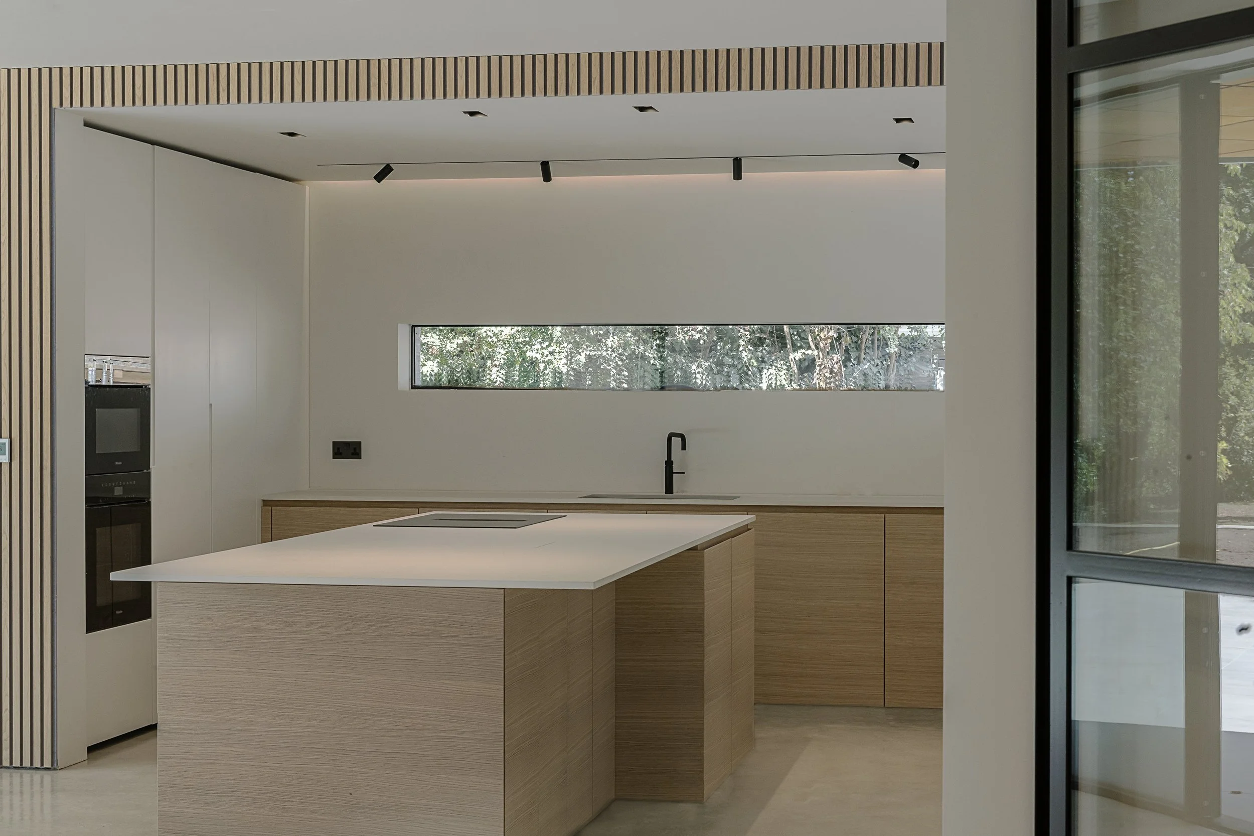 Modern kitchen with minimalist design, featuring a wooden island with a white countertop, a narrow horizontal window with green foliage outside, built-in appliances, black faucet, and floor-to-ceiling glass wall.