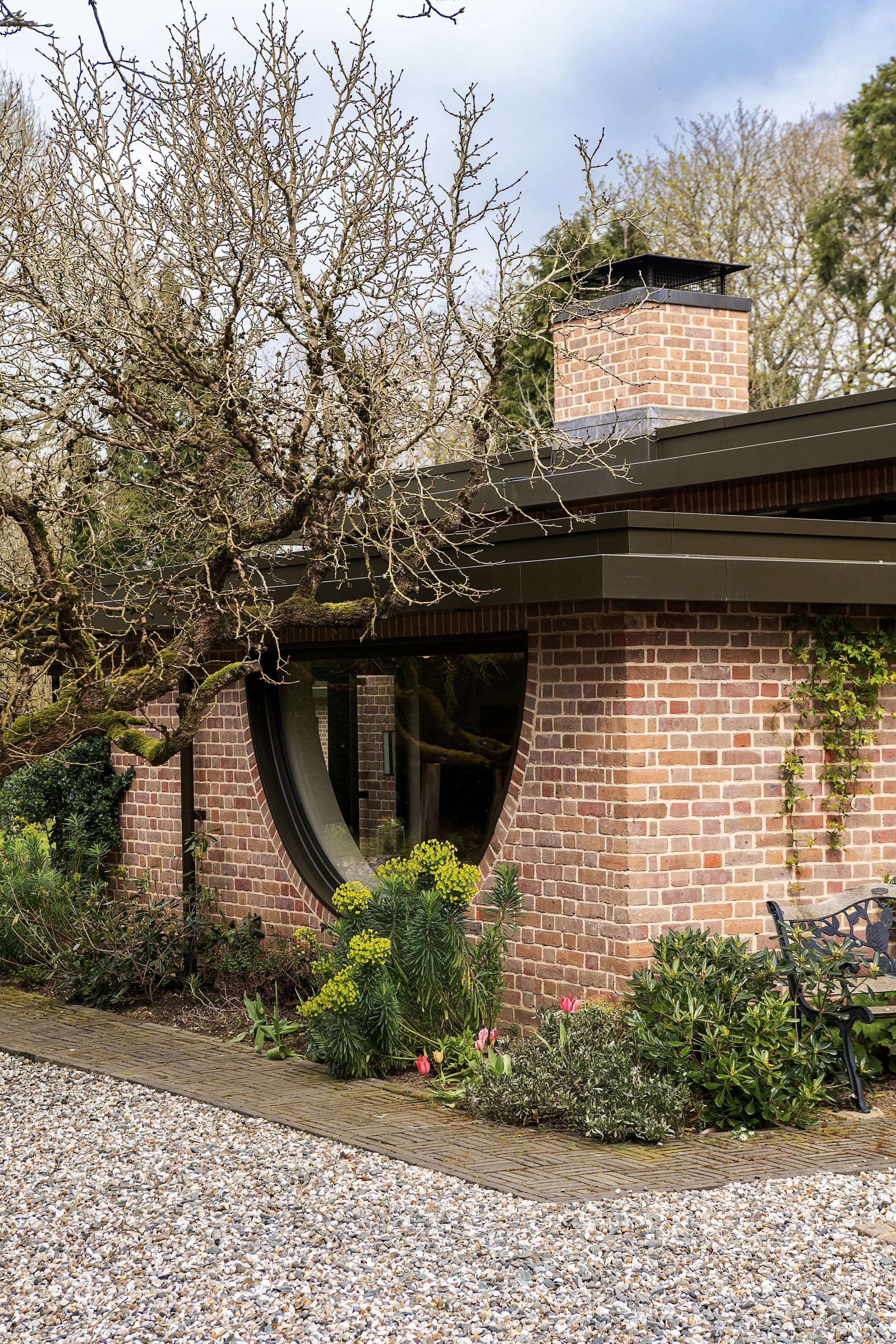 A house with a brick exterior, modern design, and a window shaped like a circle. There are plants and flowers in front of the house, and a leafless tree to the left.