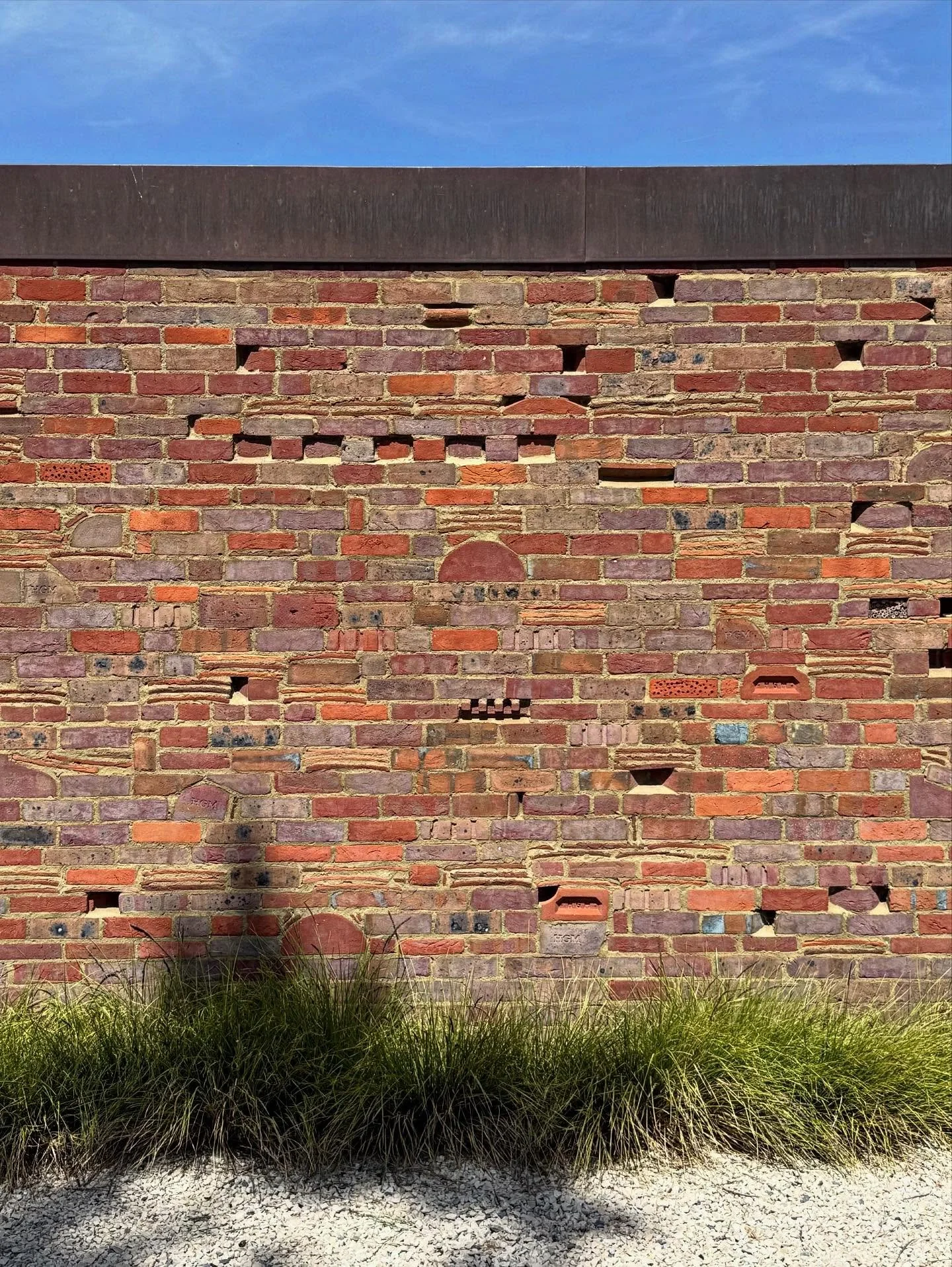A brick wall with various missing and damaged bricks, some bricks are discolored or altered, with a brown coping at the top and a clear blue sky above. Tall grass is at the base of the wall.