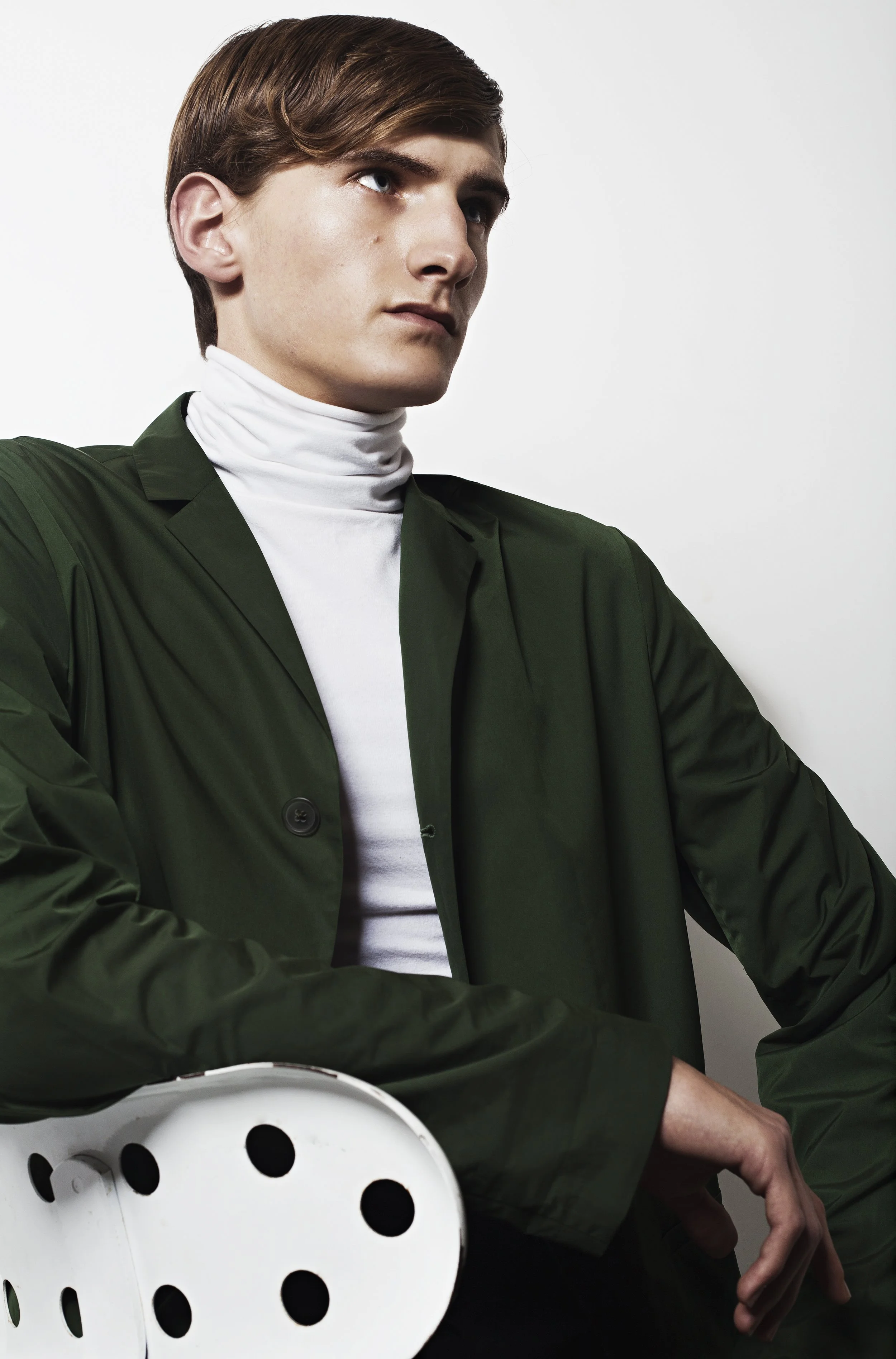 Young man with short brown hair, wearing a dark green jacket over a white turtleneck, sitting on a white chair with large circular holes, against a plain white background.