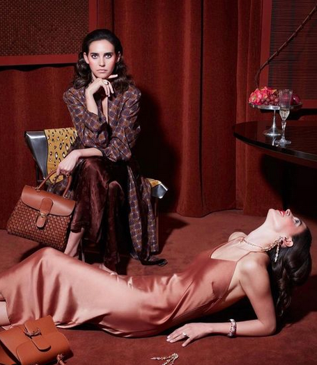 A woman in a satin gown lying on the floor, and another woman sitting in a chair with a purse, in a room with red curtains and a table with a flower arrangement.