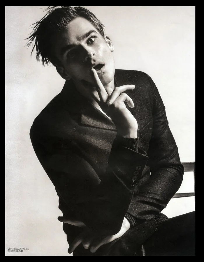 Black and white photo of a young man with styled hair, wearing a dark blazer, sitting by a wall with a hand on his face in a contemplative pose.