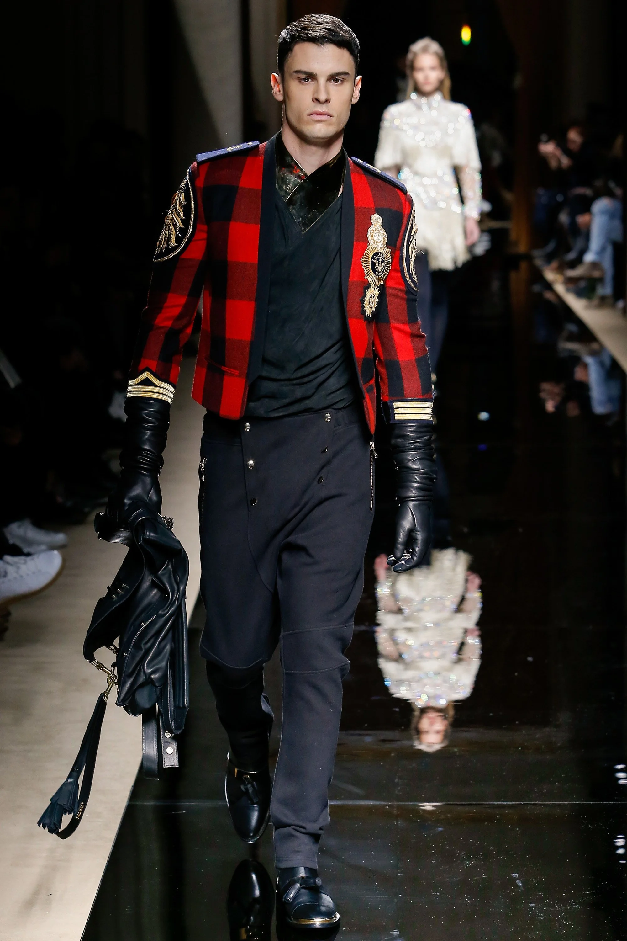 Male model walking on runway wearing a red and black plaid jacket with embellishments, black gloves, black pants, and black shoes, carrying a black jacket.