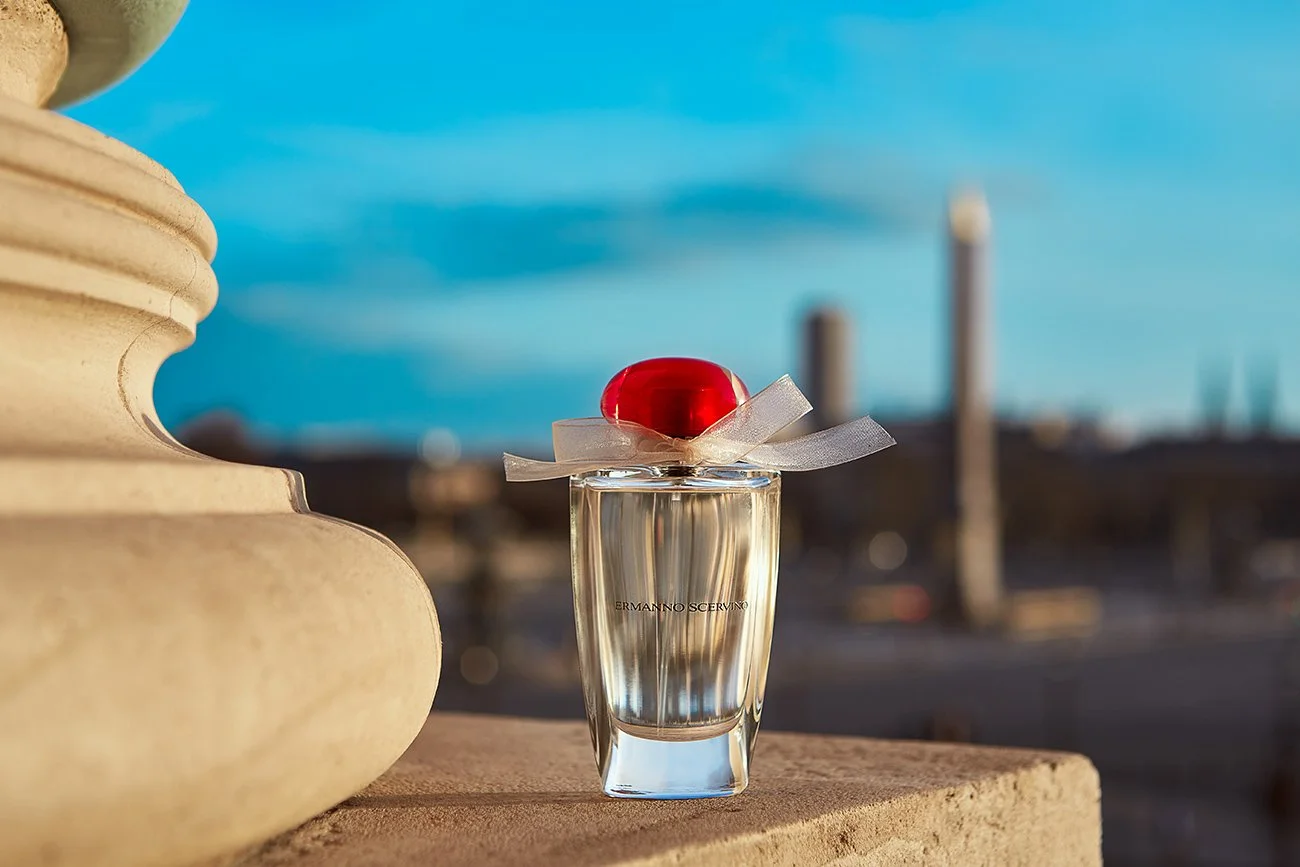 A bottle of perfume with a red lid and light-colored ribbon on top, placed on a stone ledge with a blurred cityscape and blue sky in the background.