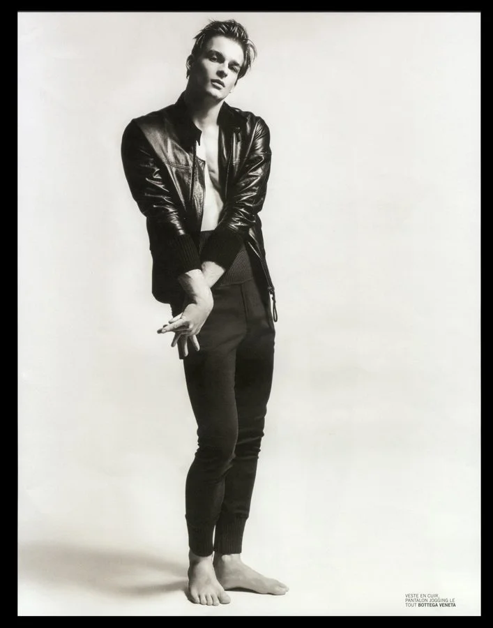 Black and white photo of a young man standing barefoot against a plain backdrop. He is wearing a leather jacket, a shirt underneath, and jogger pants. His left hand is holding something, and he is looking at the camera with a relaxed pose.