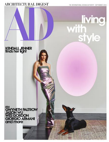 Magazine cover of Architectural Digest featuring a woman in a shiny metallic dress standing on a hardwood floor with a black and tan dog sitting next to her, and a large pink oval shape in the background.
