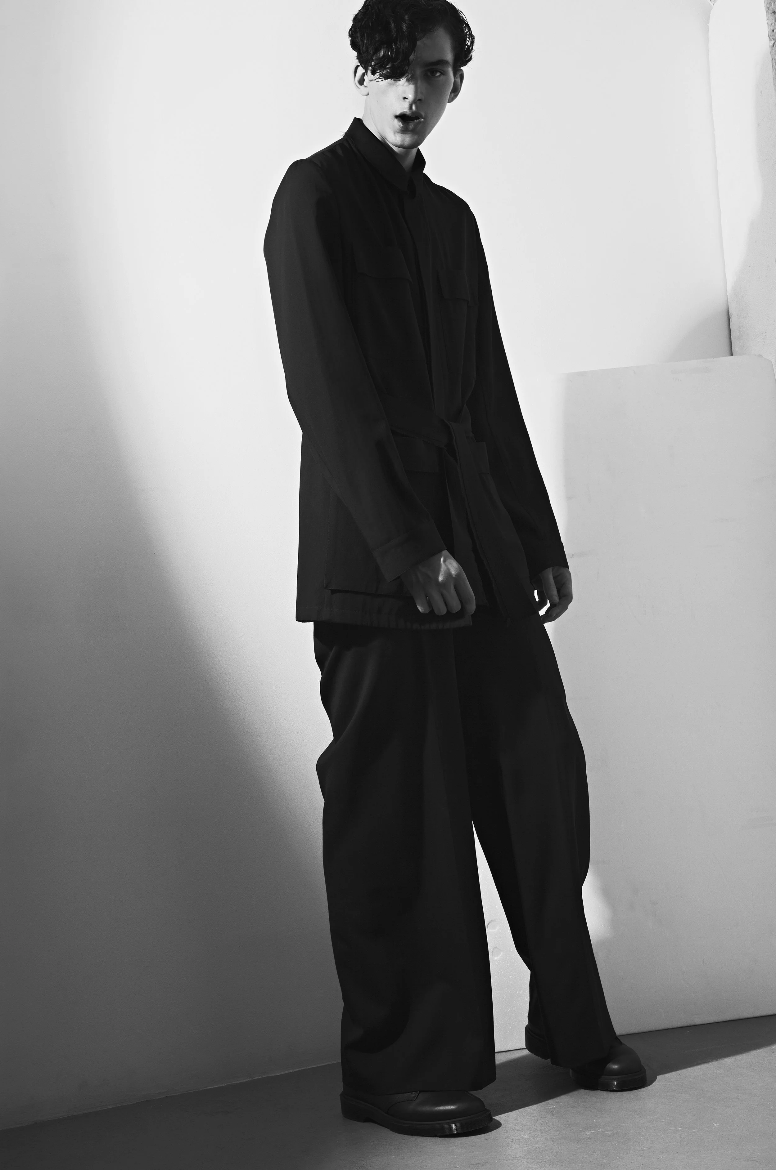 Black and white photo of a young man with dark, curly hair, wearing a long dark coat, loose pants, and boots, standing against a plain wall with a shadow cast behind him.