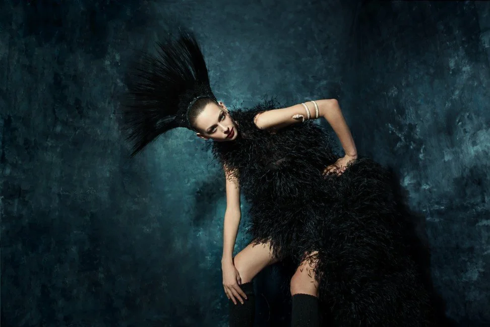 Fashion model with feathered dress, dramatic hairstyle, and jewelry posing against a textured dark background.