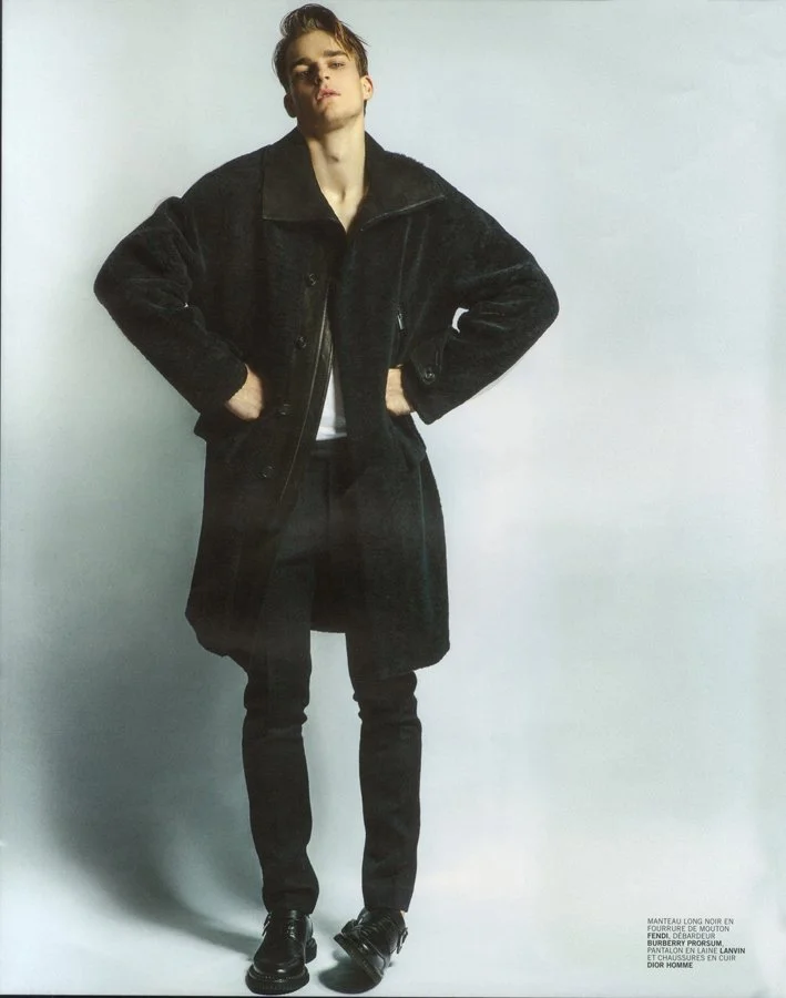 A young man standing with hands on hips wearing a long black coat, black pants, black shoes, and a white shirt underneath, posing against a plain light-colored background.