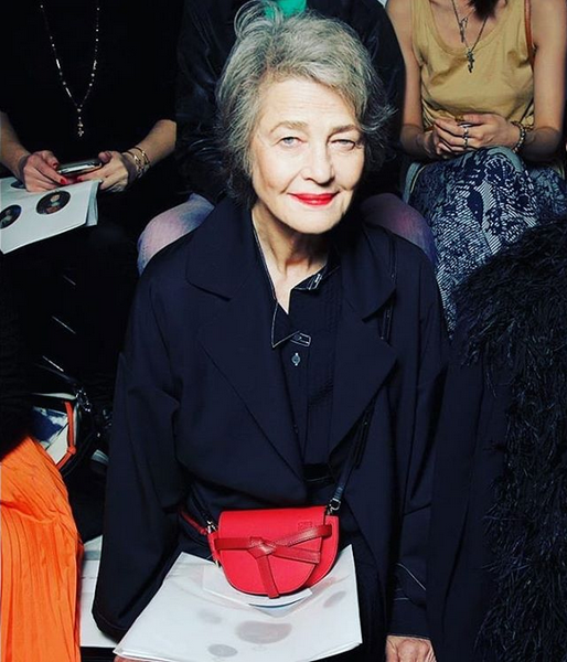An elderly woman with short gray hair smiling at the camera, wearing a navy blue jacket and a red crossbody bag, seated among other people.