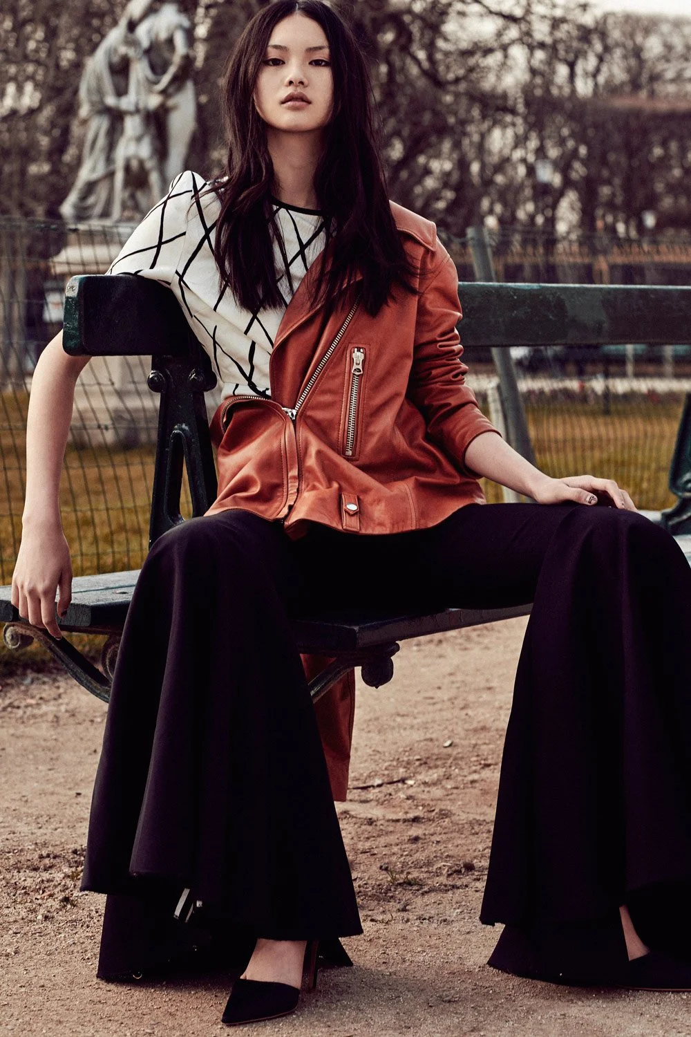 Young woman sitting on a park bench, wearing a white T-shirt with black geometric lines, an orange leather jacket draped over her shoulders, wide black trousers, and black high-heeled shoes.