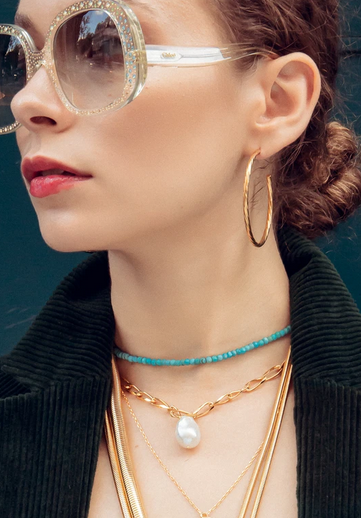 Close-up of a woman wearing large sunglasses, hoop earrings, multiple layered necklaces including a pearl pendant, and a black jacket.