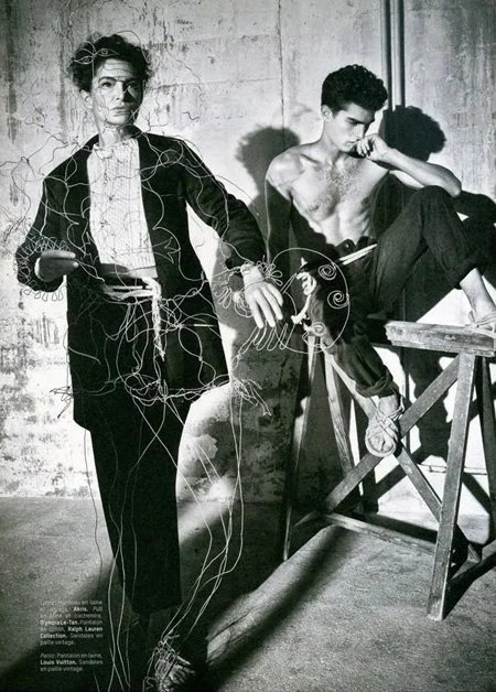 Black and white photo of a woman with glasses and messy hair holding a cell phone, and a shirtless man with curly hair sitting on a wooden platform, both surrounded by tangled string or wires, against a textured wall.