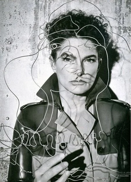 A woman with curly hair wearing a leather jacket and a shirt, holding a cigarette, with wire art that outlines her face and head in front of a concrete wall.