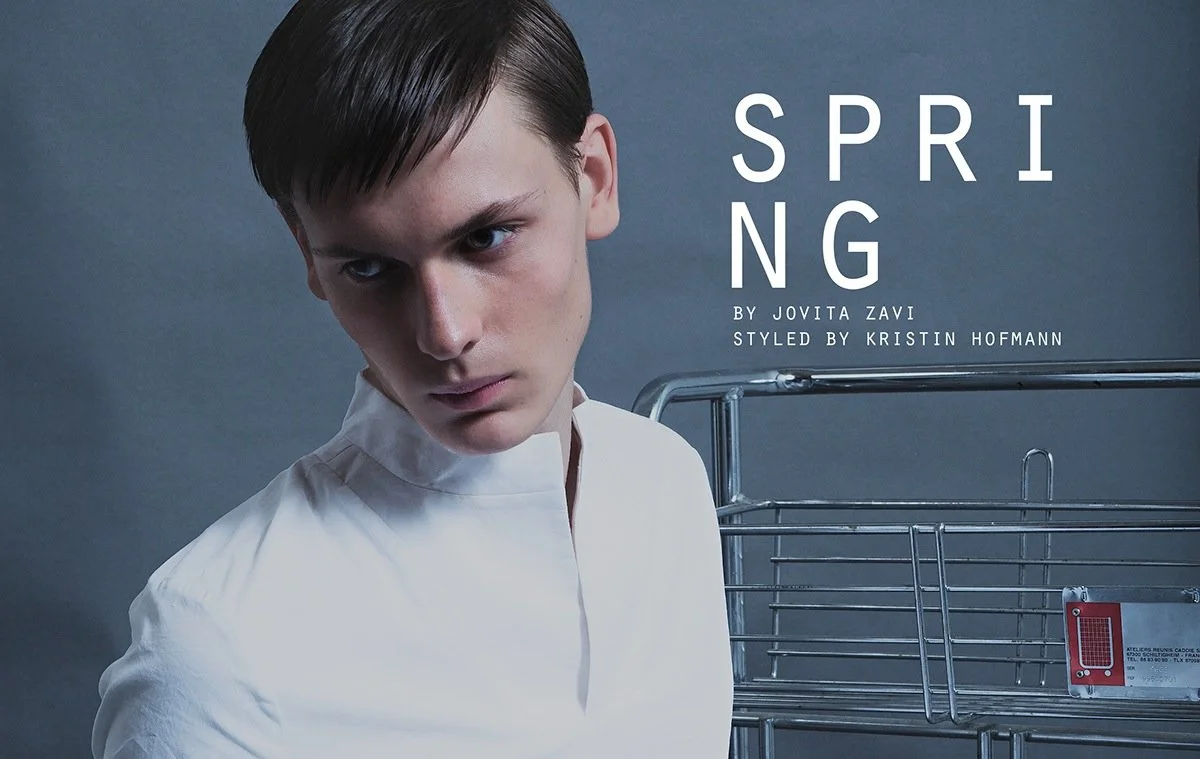 Young person with short dark hair looking at camera, standing next to a metal shopping cart, against a plain background.