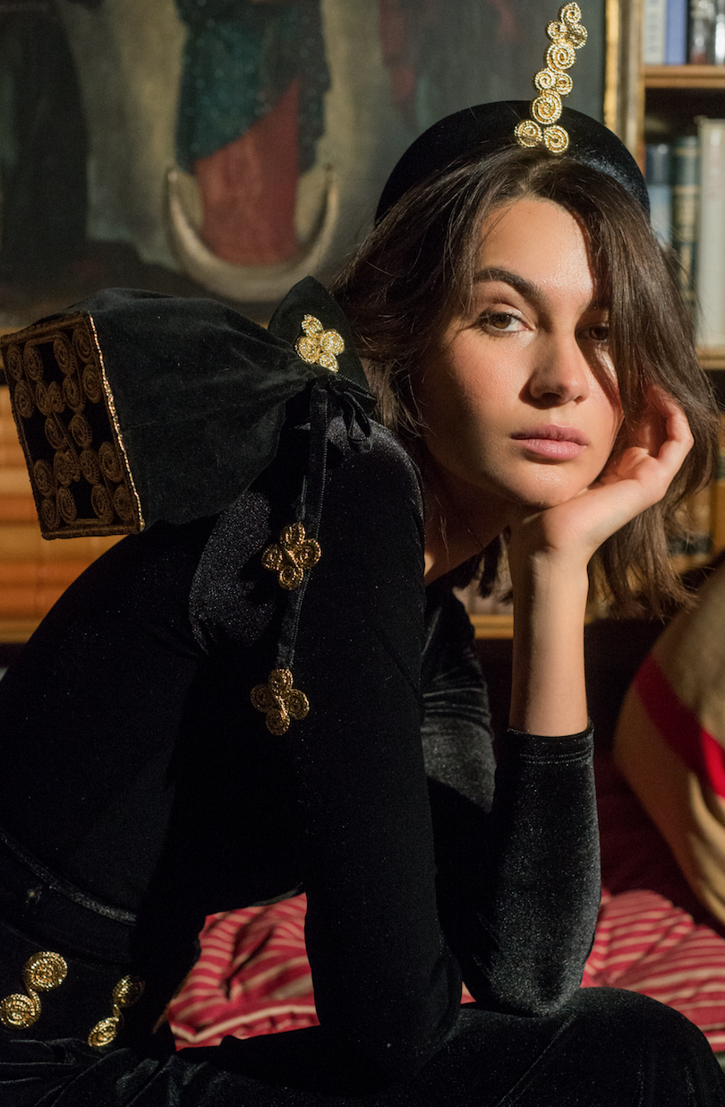 Young woman with wavy brown hair, wearing a black velvet dress with gold embellishments, sitting with her chin resting on her hand in an indoor setting with a painting and bookshelf in the background.