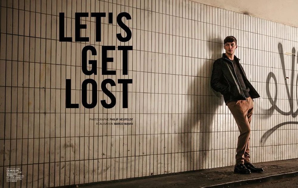 Young man leaning against a tiled wall with graffiti, standing next to large black text that reads "Let's Get Lost."