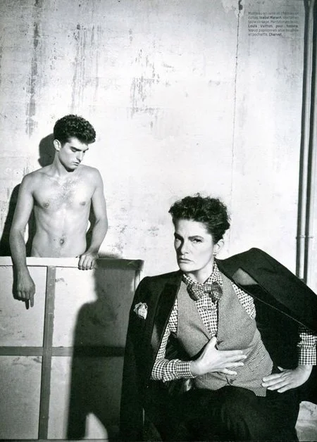 Black and white photo of a woman sitting with her hand on her stomach, wearing a patterned shirt and blazer, with a serious expression, and a young shirtless man standing behind a barrier in a stark, industrial setting.