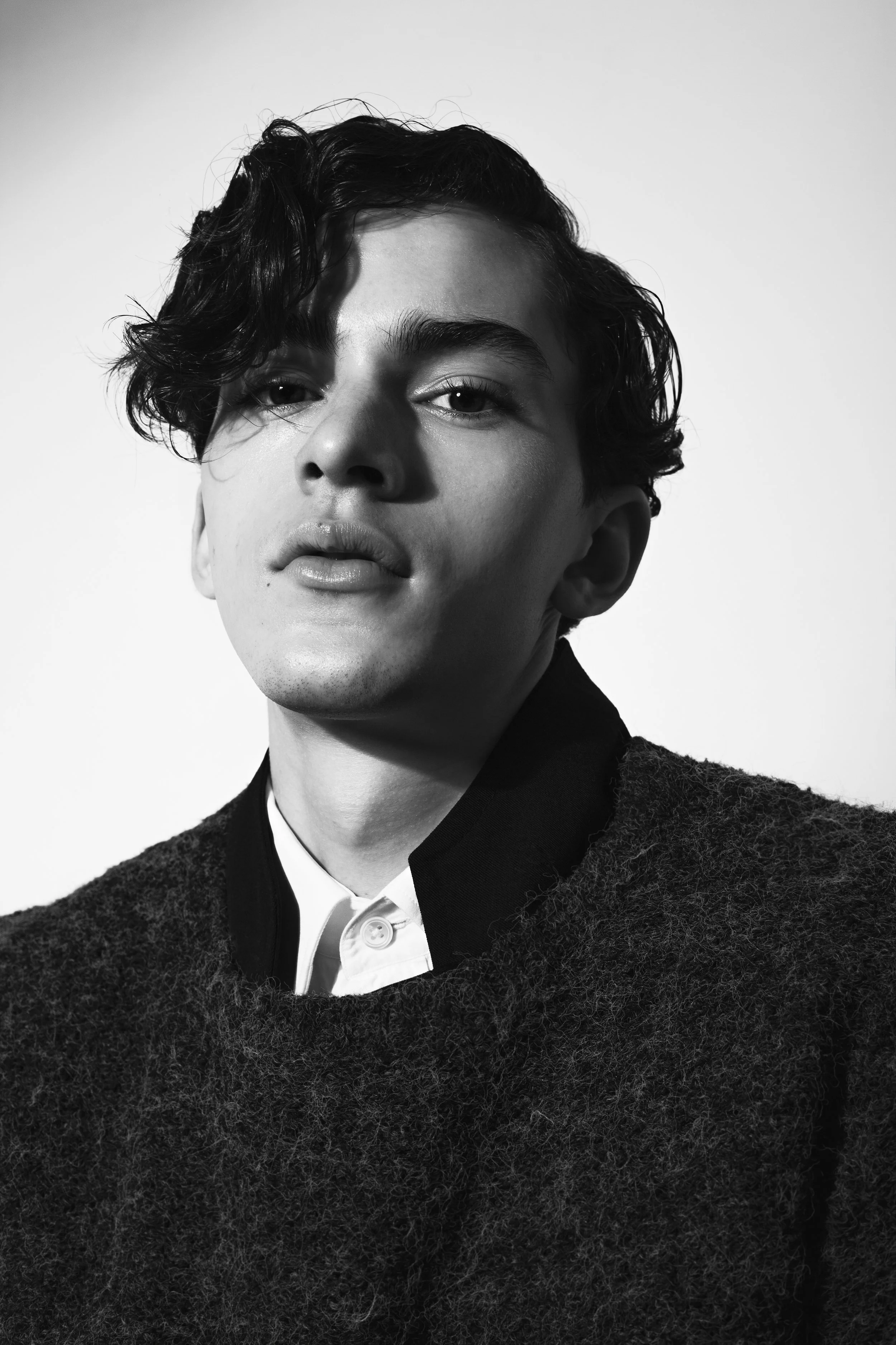Black and white portrait of a young person with wavy hair, wearing a white shirt and a dark textured sweater, looking confidently at the camera.
