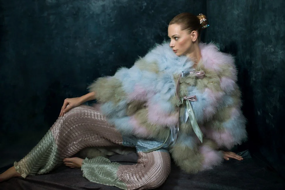 A woman sitting against a dark backdrop, wearing a pastel-colored fur coat and patterned pants.