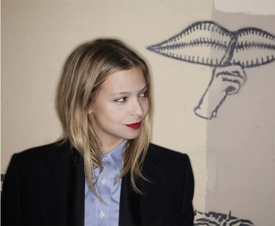 A woman with blonde hair, red lipstick, wearing a black blazer over a light blue shirt, standing near a wall with a drawing of lips and a nose.