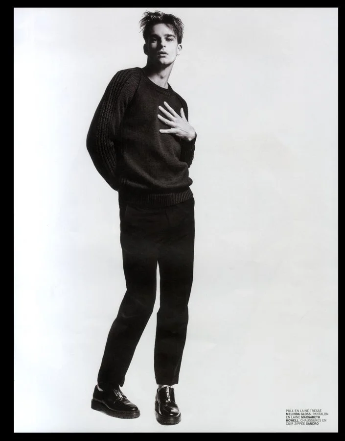 Black and white fashion photograph of a young man standing against a white background, wearing a dark sweater and dark pants, with one hand on his chest and looking at the camera.