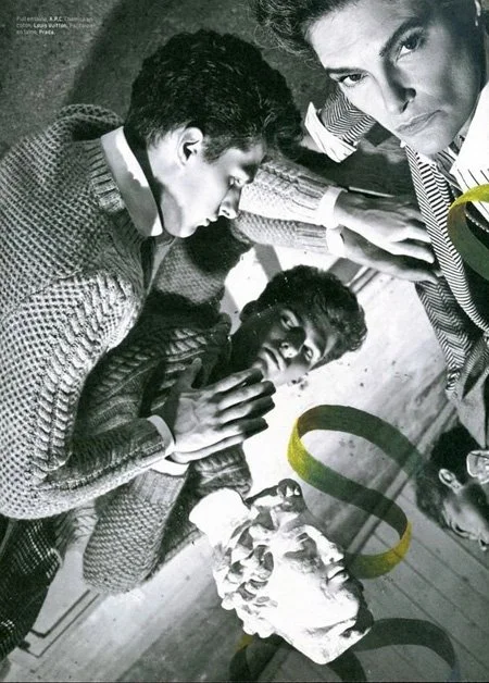 Black and white photo of three young men closely gathered around a cake, with one looking directly at the camera, all appearing to be in a playful or celebratory moment.