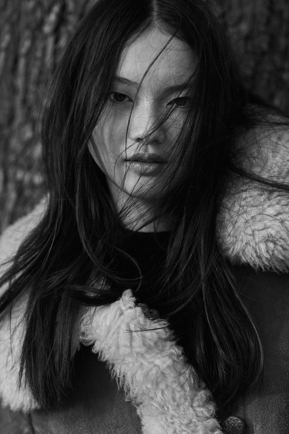 Black and white portrait of a woman with long hair, partially covering her face, wearing a coat with a shearling collar.