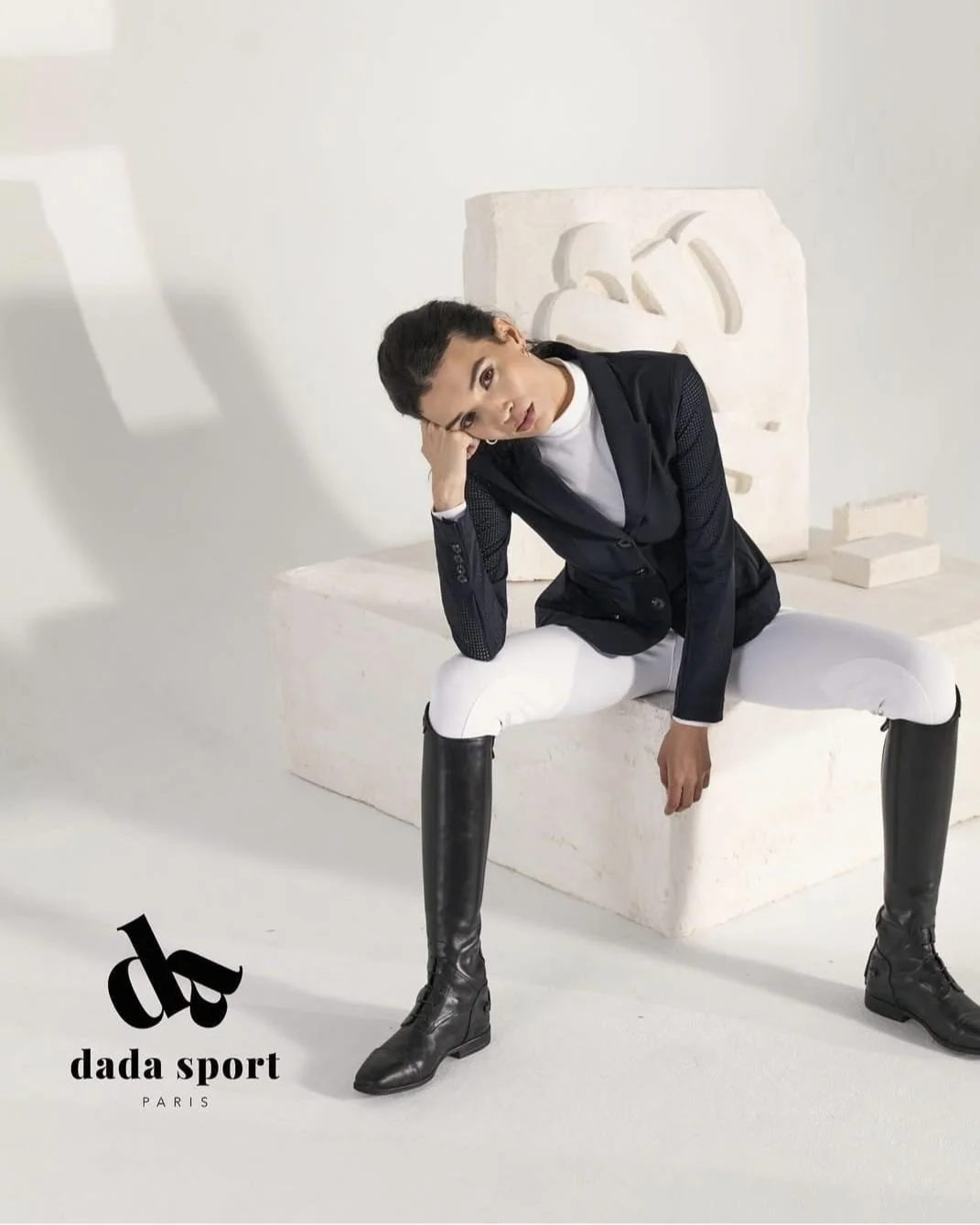 Model sitting on a white block, wearing a black blazer, white pants, and black boots, with a white background and a carved stone sculpture behind her. DADA SPORT PARIS logo in the bottom left corner.