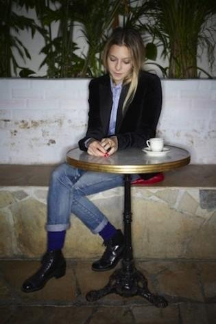 A young woman sitting at a small round table with a white coffee cup and saucer, looking down at her phone, in a space with plants and a white brick wall.