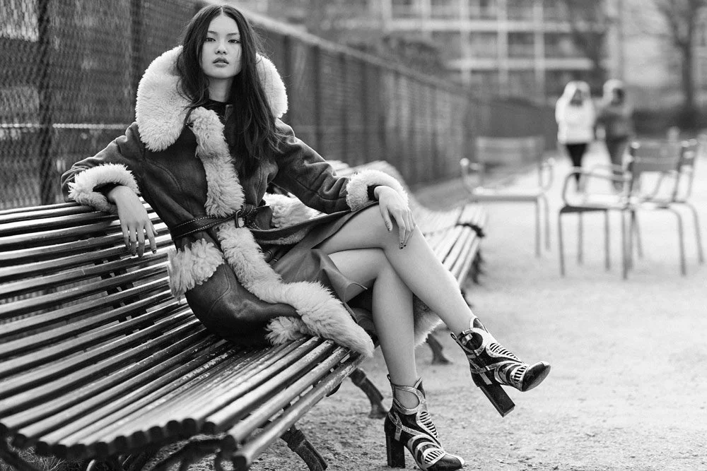 A young woman with long dark hair sitting on a park bench wearing a shearling-lined coat and patterned ankle boots, with a fence and other park benches in the background.