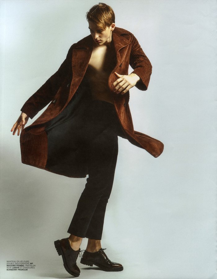 A person in mid-movement wearing a brown coat, black pants, and black shoes, against a plain light background.