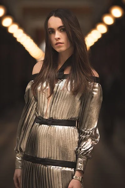 Young woman with long dark hair wearing a metallic gold and black dress, standing in a dimly lit corridor with blurred lights in the background.