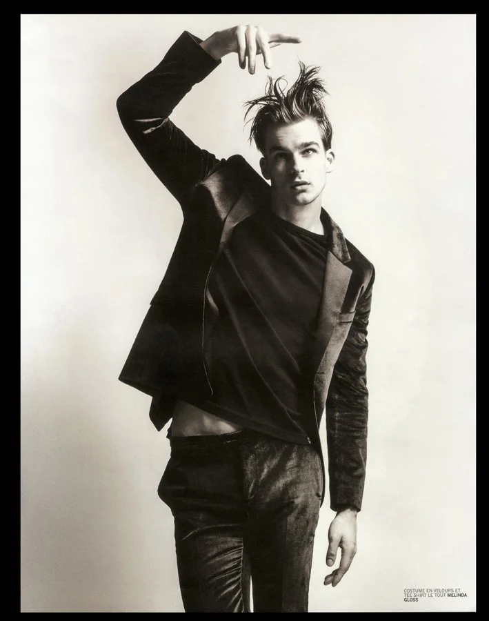 Black and white photo of a young man with messy hair wearing a dark jacket and pants, posing with one arm raised and pointing downward, against a plain light background.