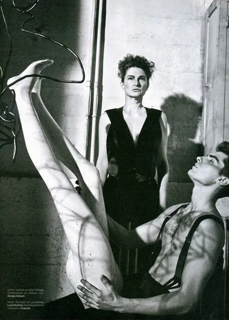 Black and white photo of two women, one standing and one sitting. The standing woman is wearing a sleeveless top and has curly hair, looking into the distance. The sitting woman is leaning back with her legs raised, wearing a tank top and shorts, loo