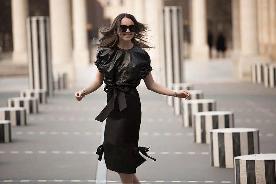 Woman in black leather top and skirt, wearing sunglasses, smiling with hair flowing, standing outdoors among striped bollards.