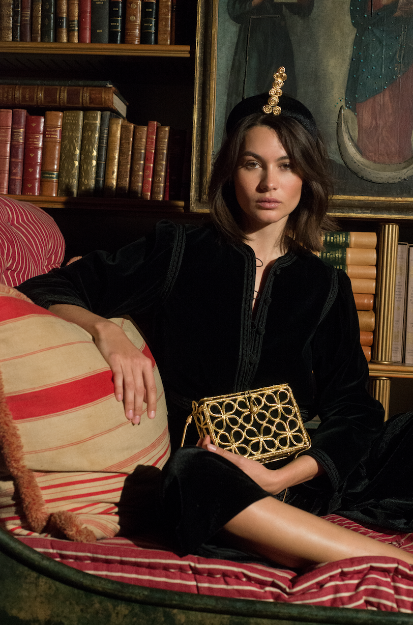 A woman with shoulder-length brown hair sitting on a striped pillow in a dimly lit room, holding a gold decorative clutch. She wears a black velvet dress or coat and a headband with a gold ornament. In the background, there are bookshelves filled wit