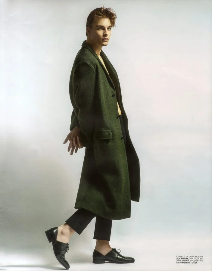 A young person with styled hair wearing a long green coat, black pants, and black shoes standing against a plain background.