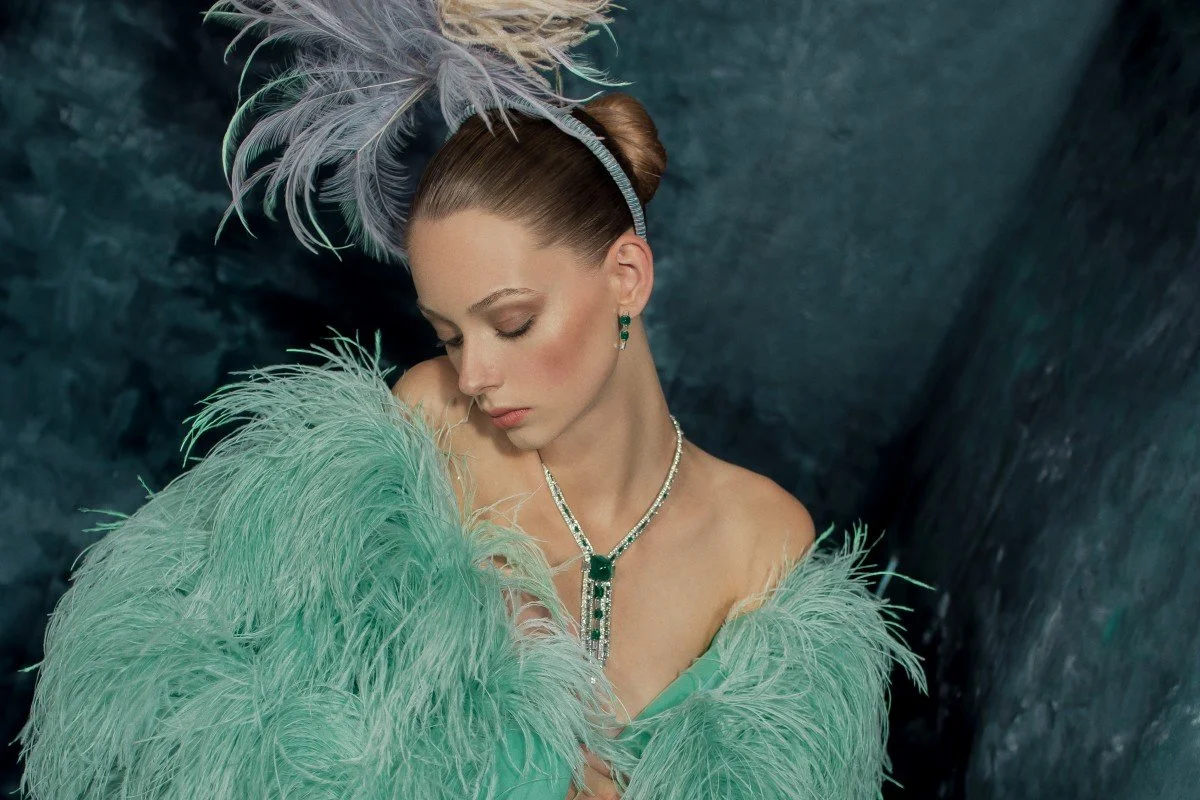 A woman dressed in a glamorous turquoise feathered gown, wearing matching jewelry and a feathered headband, with her eyes closed and head tilted down.