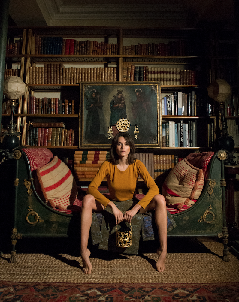A young woman with shoulder-length brown hair, wearing a mustard yellow top and a patterned skirt, sits barefoot on an ornate vintage-style sofa with striped cushions. She holds a decorative lantern in her hands. Behind her is a large wooden bookshel