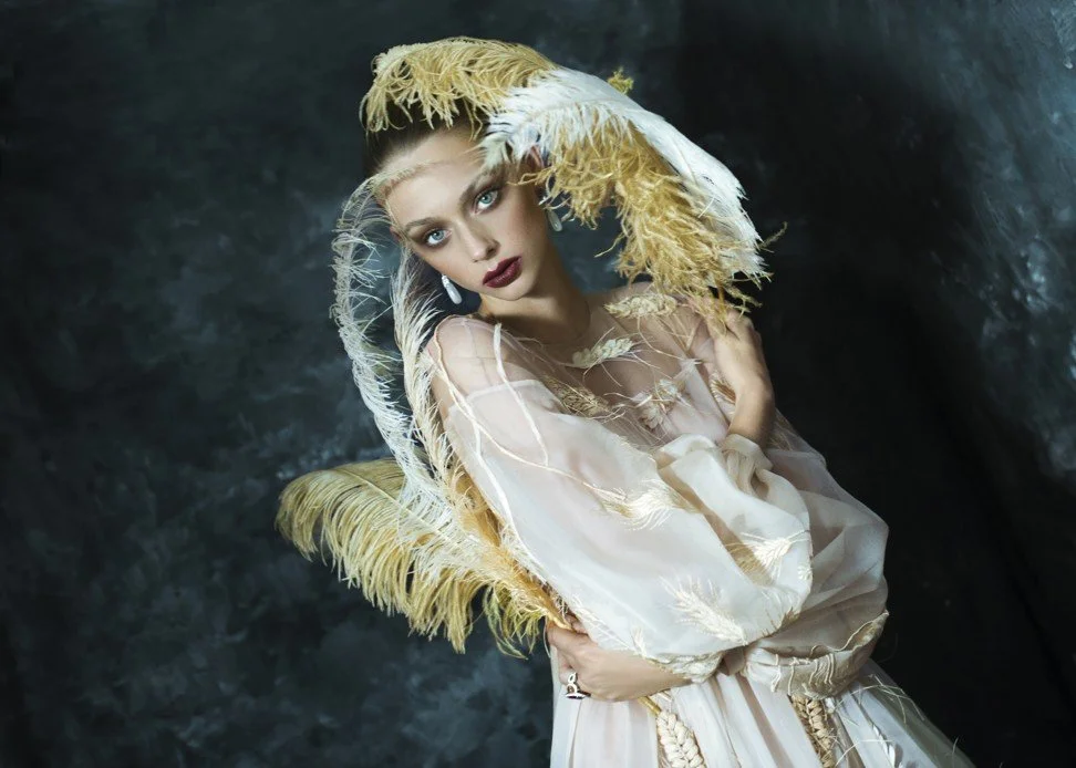 A woman with dramatic makeup and blonde hair, wearing an elaborate cream-colored dress and a large feathered hat, standing against a dark textured background.