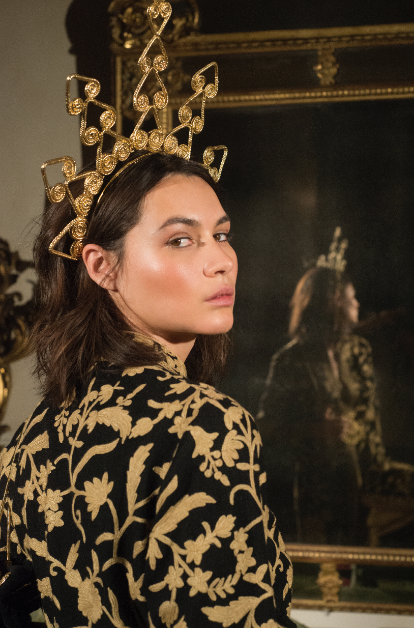 A woman with dark brown hair wearing a gold crown and black and gold embroidered clothing, standing in front of a mirror that reflects her image and a dark background with ornate gold detailing.