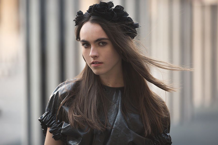 A young woman with long brown hair wearing a black floral headband and black leather top, standing outdoors with a blurred background.