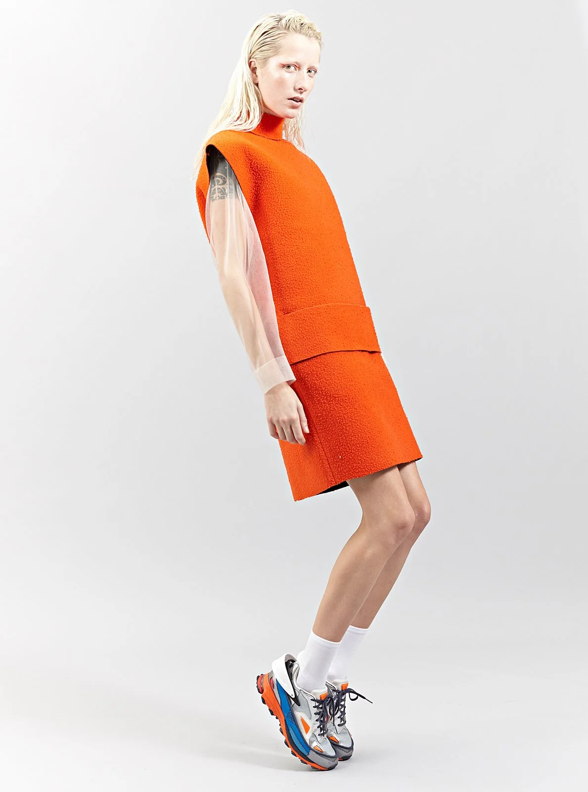 A woman with long, bleached blonde hair poses in a bright orange outfit consisting of a sleeveless top and a matching skirt. She is wearing white socks and colorful athletic sneakers, standing against a plain white background.