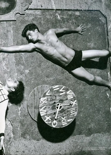 Black and white photo of a shirtless man lying on the ground on a yoga mat with a clock in front of him, and a woman lying next to him, both on concrete surface.