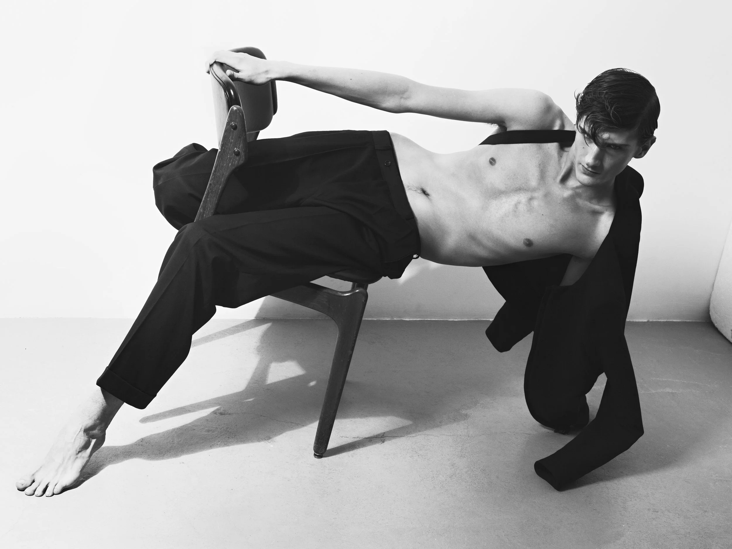Black and white photo of a shirtless young man with dark hair, wearing black pants and a blazer, posing with one hand on a wooden chair and the other on the floor, in a minimalist room with plain wall and carpet.