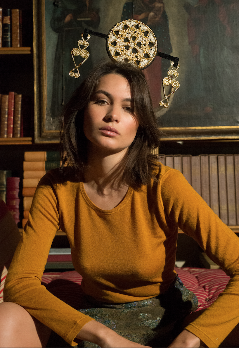 A young woman sitting in a room with a dark background, wearing a mustard yellow long-sleeve shirt and an elaborate gold headdress with intricate designs. There are books and framed artwork behind her.