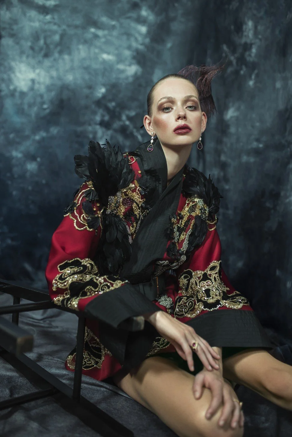 A woman dressed in a luxurious, ornate garment with black, red, and gold embroidery, sitting on a black chair against a dark, textured background. She has makeup, earrings, and a hairstyle with a swept-up design.