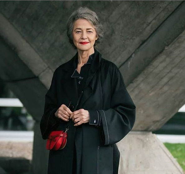 Older woman with short gray hair, wearing a black coat, standing outdoors in front of a concrete structure, holding a small red purse.